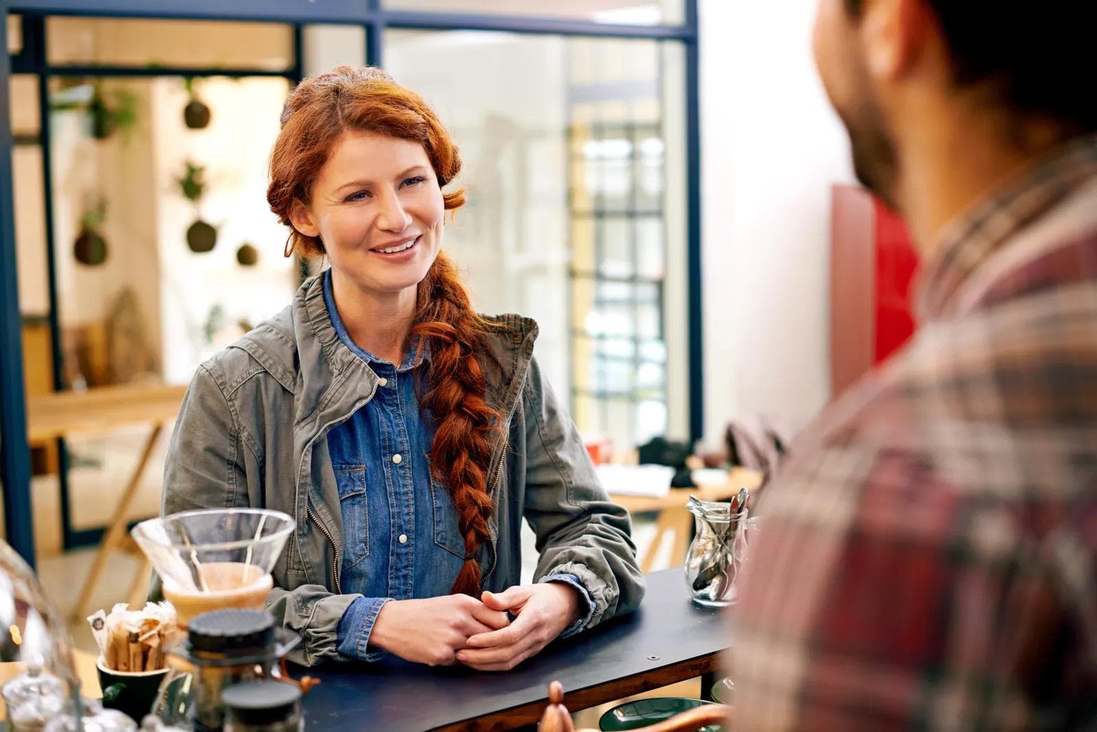 A woman with a long red braid leaning on a café counter, smiling as she talks with the person across from her