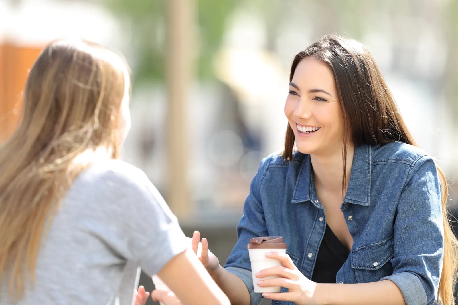 Two young women having an animated conversation outdoors, one smiling and holding a takeaway coffee cup